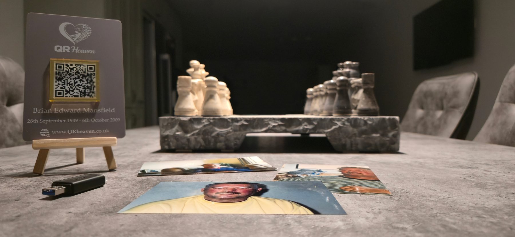 A QR Heaven plaque standing on a family table beside printed photographs and a chess set